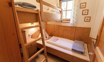 A cozy room with a loft bed structure and a single bed. The walls are made of wood, and there are large windows that brighten the space.