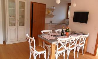 A modern dining room with a large wooden table and white chairs. In the background, a kitchen and a television can be seen.