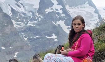 A girl sits on a meadow in the mountains and observes two marmots. Snow-covered peaks can be seen in the background.