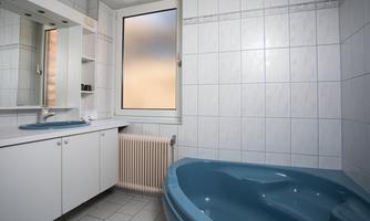 A modern bathroom with a blue bathtub and large tiles. The window provides natural light and a bright atmosphere.