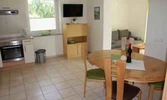 A bright, modern kitchen with a wooden dining area. In the background, a small living area with a gray sofa is visible.
