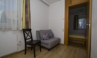 A cozy room with a gray sofa and a black chair. In the background, a bedroom can be seen.