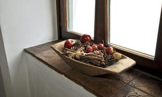 A wooden bowl with red fruit and several cinnamon sticks is on a windowsill. In the background are large windows that let in a lot of light.