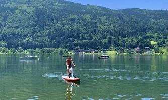 A man is standing on a paddleboard on a calm lake. The surroundings are green with hills and boats in the background.