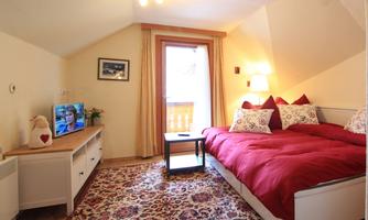 A cozy bedroom with a large bed and red bedspreads. A television is on a white cabinet, and there is a balcony with a view outside.