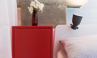 A modern bedroom with a white bed and a red nightstand. On the nightstand, there is a vase with white flowers.