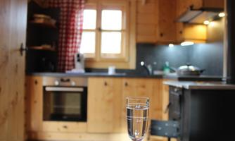 A cozy kitchen with wooden shelves and a window that lets in daylight. In the foreground, there is a glass of water on the table.