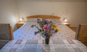 A cozy bedroom with a bed and a tablecloth. In the middle is a vase with colorful, dried flowers.