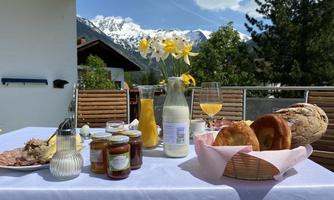 A set table with fresh bread, jam, and milk in front of a picturesque mountain landscape. In the background, snow-capped mountains and a blue sky can be seen.