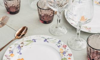 A beautifully set table with floral patterns on the plates and elegant glasses. The table is outdoors and surrounded by green grass.