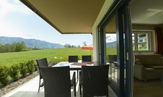 A cozy terrace with a table and chairs, surrounded by green meadows. In the background, mountains and a sun umbrella are visible.