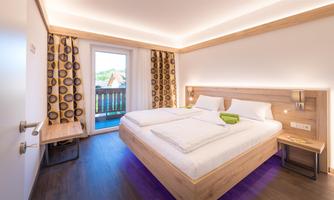 A modern hotel room with two single beds and wooden furniture. Large windows provide plenty of natural light and access to a balcony.