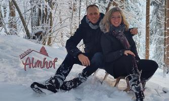 A couple is sitting in the snow and smiling. In the background, there is a sign with the inscription "Almhof."