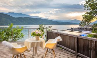 A beautiful balcony with a view of a lake and mountains in the background. A table with flowers and two chairs invites you to relax.