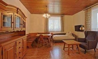 A cozy living room with wooden paneling and a large dining table. There is a sofa, an armchair, and plenty of natural light coming through the windows.