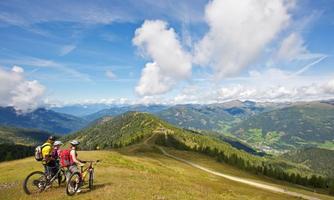 Two cyclists are standing on a hill and enjoying the view of the valley. The sky is blue with some clouds and the landscape is green and mountainous.