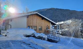 A modern house with wooden cladding stands in the snow. In the background, snow-covered mountains and a clear blue sky can be seen.