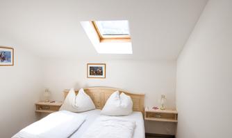 A bright bedroom with two separate beds and a skylight. Pictures hang on the walls, and there are small nightstands with lamps.