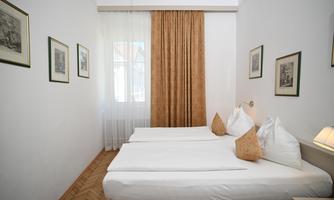 A simple, bright room with two single beds and cozy pillows. Pictures hang on the walls, and the room has large windows with curtains.