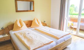 A cozy bedroom with two single beds and bright pillows. Large windows offer a view of the balcony and the green surroundings.