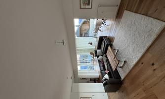 A modern living room with a black sofa and a wooden table. Large windows offer a view of the surroundings and let in a lot of light.