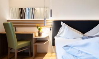 A modern hotel room with a cozy bed and a desk. On the table, there is a flower in a pot and there is a large mirror on the wall.