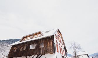 A picturesque wooden house in the snow with a steep roof. In the background, gentle hills and a cloudy sky can be seen.