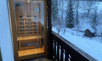 A wooden porch with a glass box that houses a sauna. In the background, snow-covered trees and a small cottage can be seen.