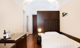A modern room with a double bed and wooden floor. There is a nightstand and a chest of drawers with bottles and a glass.