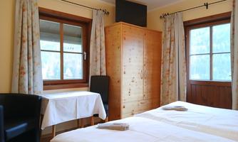 A cozy room with two single beds, a table, and a chair. Large windows let in plenty of daylight, and the room is designed to be bright and welcoming.