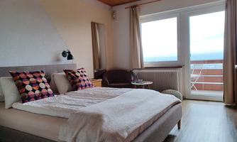 A cozy bedroom with a large bed and colorful pillows. There is a balcony with windows that offer a view outside.