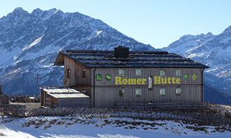 A traditional mountain hut named "Römer Hütte" in a snow-covered Alpine panorama. Majestic mountains rise in the background under a clear sky.