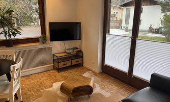 A cozy living room with a black sofa and a rug. Large windows provide a view of the outdoor area and bring in plenty of daylight.