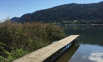 A wooden pier by the shore of a calm lake. In the background, there are wooded hills and a blue sky with a few clouds.