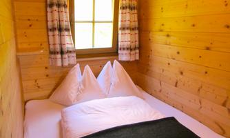 A cozy bedroom with wooden paneling and a window. The bed is furnished with pillows and a blanket.