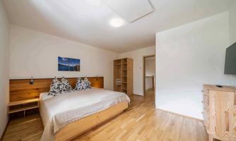 A bright bedroom with a large wooden bed and decorative pillows. There is a desk and a television on the wall.