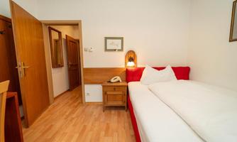 A small, cozy room with two single beds and warm wood decor. There is a door leading to a hallway and a dresser next to the bed.
