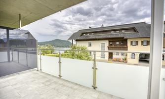 A balcony with glass railing and a view of a quiet residential building. The sky is cloudy and hills are visible in the background.