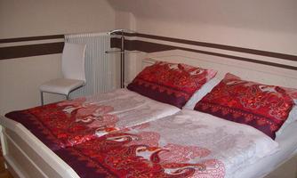 A cozy bedroom with a large bed and colorful bedcovers. Next to the bed is a white chair and a small table.