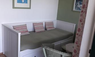 A cozy room with a white sofa bed and striped cushions. A table with a rattan chair is in the corner, and the walls are painted in soft colors.