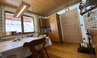 A cozy kitchen with a dining table, a wooden cupboard, and windows with curtains. The light is warm and inviting, and the walls are simply designed.