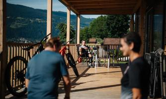 A wooden terrace with a view of the mountains. Two people are visible, one is sitting relaxed while the other is moving.