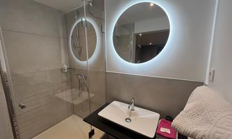 A modern bathroom with a walk-in shower and a sleek sink. Large, illuminated mirrors complete the stylish design.