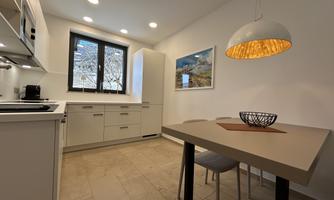 A modern kitchen with bright furniture and a square table. A large window brings plenty of daylight into the room.