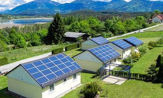 A view of several houses with solar panels in a green landscape. In the background, mountains and a body of water can be seen.