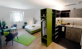 A modern room with a sleeping area and a kitchenette. The decor is in green and neutral colors and looks inviting.