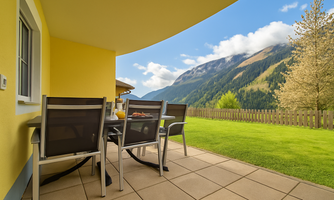 A terrace with modern furniture and a view of green hills and mountains. The sky is blue with some clouds.