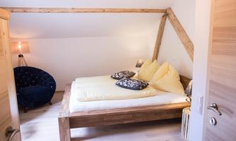 A cozy bedroom with a large bed, yellow cushions, and a blue armchair. The rustic wooden beams give the room a warm character.