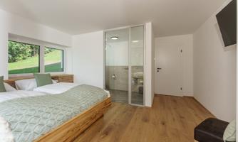 A modern bedroom with a double bed and wooden furniture. In the background, there is a bathroom with a glass wall.