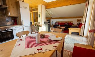 A cozy kitchen with a wooden table and modern furniture. In the background, a living room and a bright atmosphere can be seen.